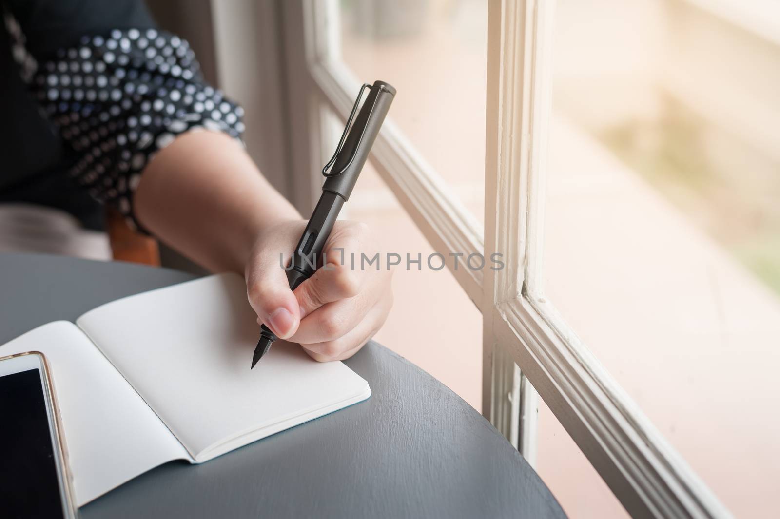 Left-handed woman holding pen while writing on small notebook beside window. Freelance journalist working at home concept.