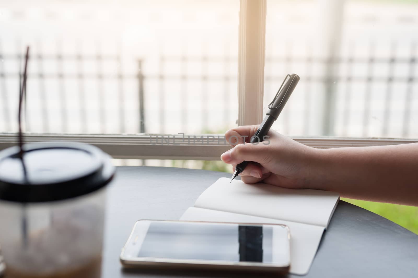 Woman hand holding pen while writing on small notebook with smartphone on table beside window. Freelance journalist working at home concept.