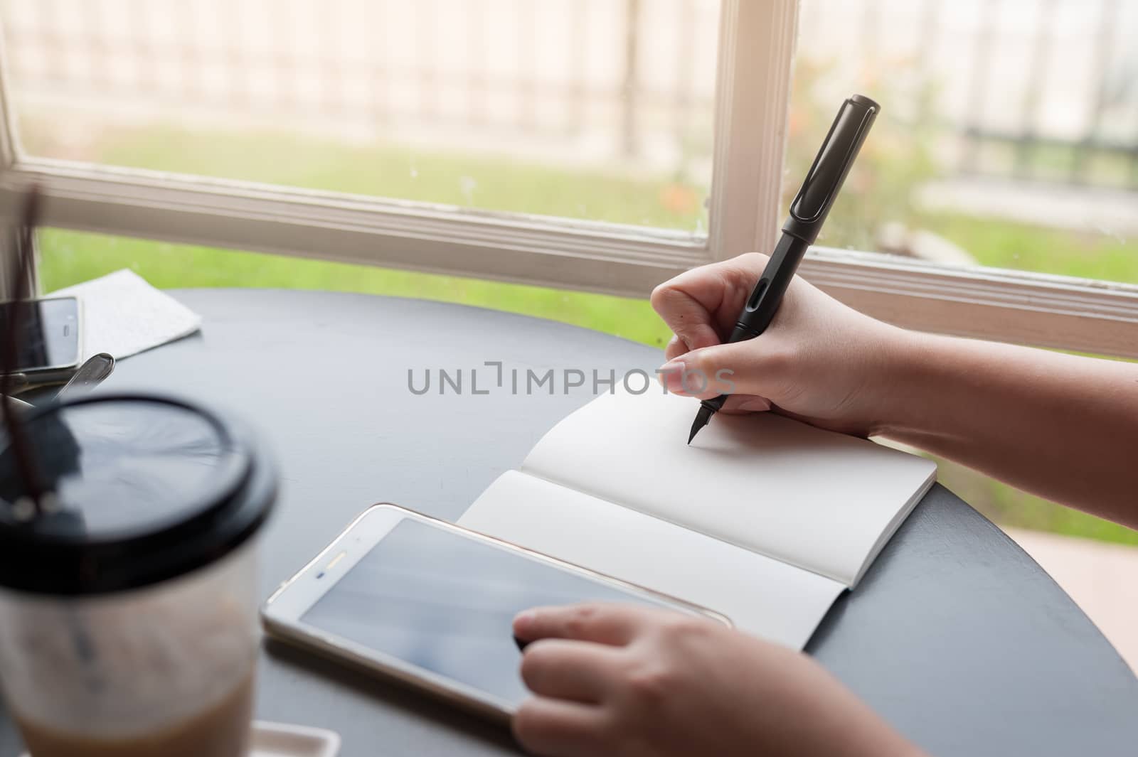 Woman hand writing a note on notebook while another hand touching on smartphone screen on table beside window. Working from anywhere concept