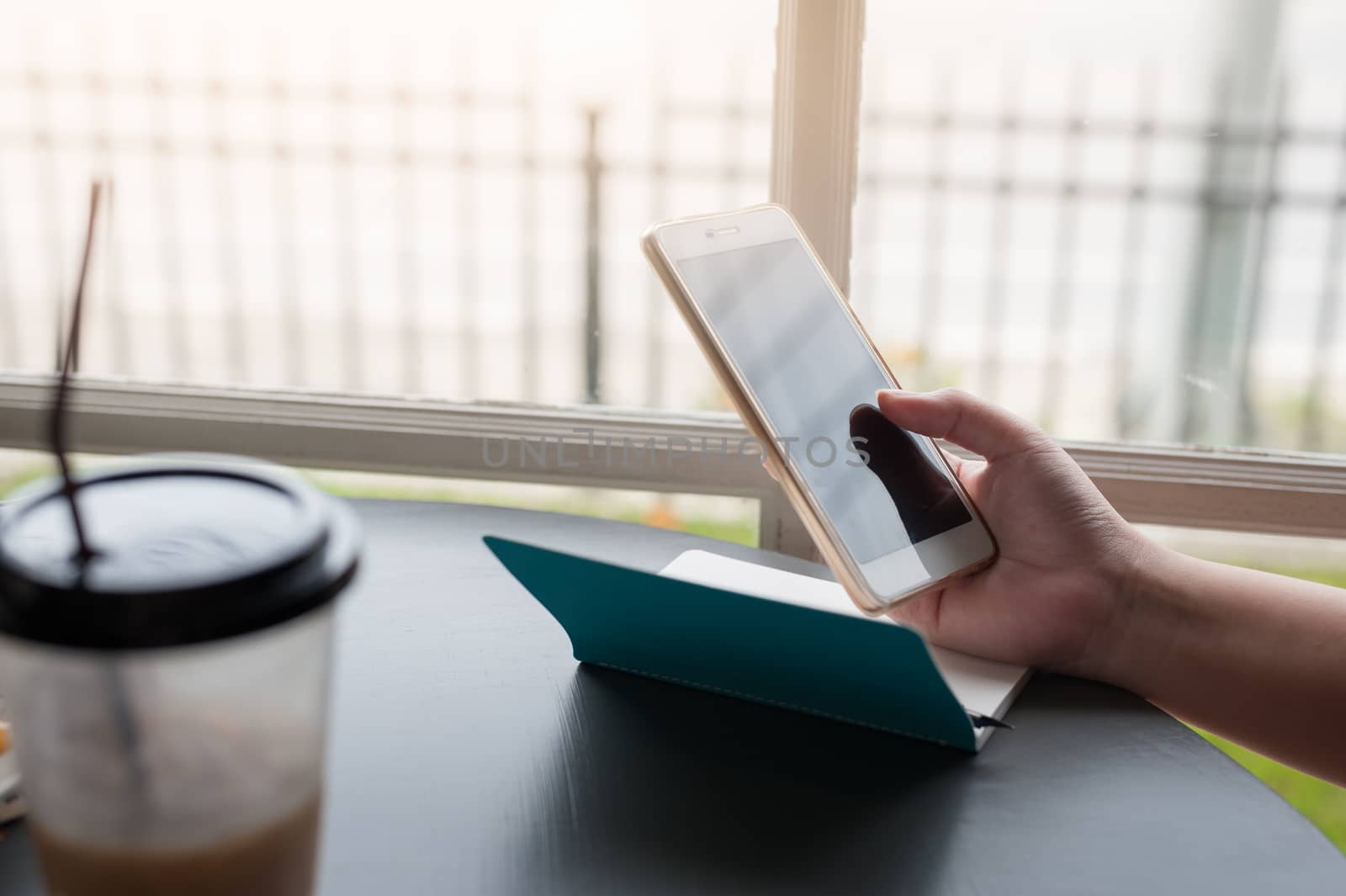 Woman hand holding smartphone beside window with coffee cup and small notebook on table