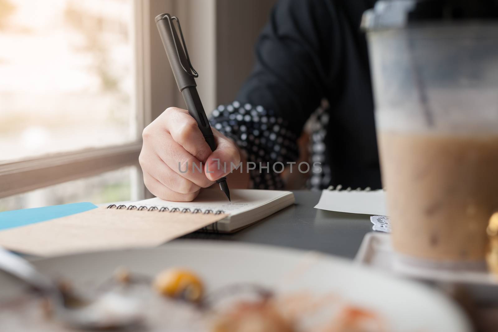 Woman hand holding pen while writing on small notebook beside window. Freelance journalist working at home concept.