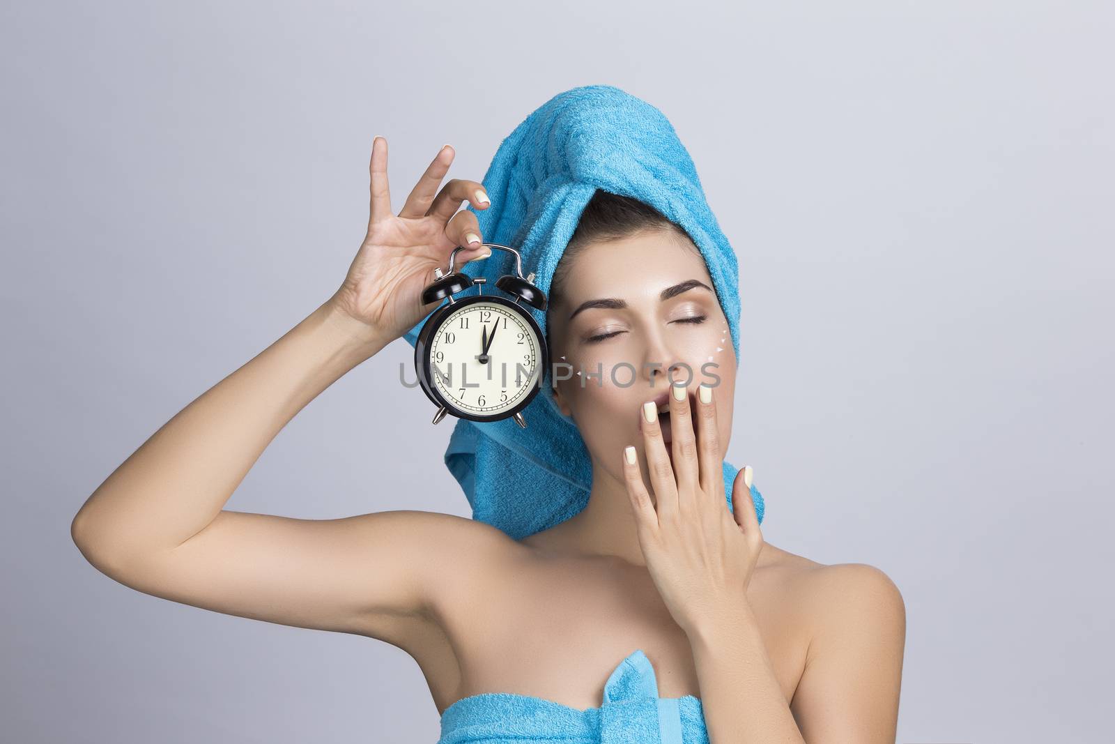 Tired yawning woman in towel on head and creme on face holding alarm clock showing midnight, night creme skincare concept