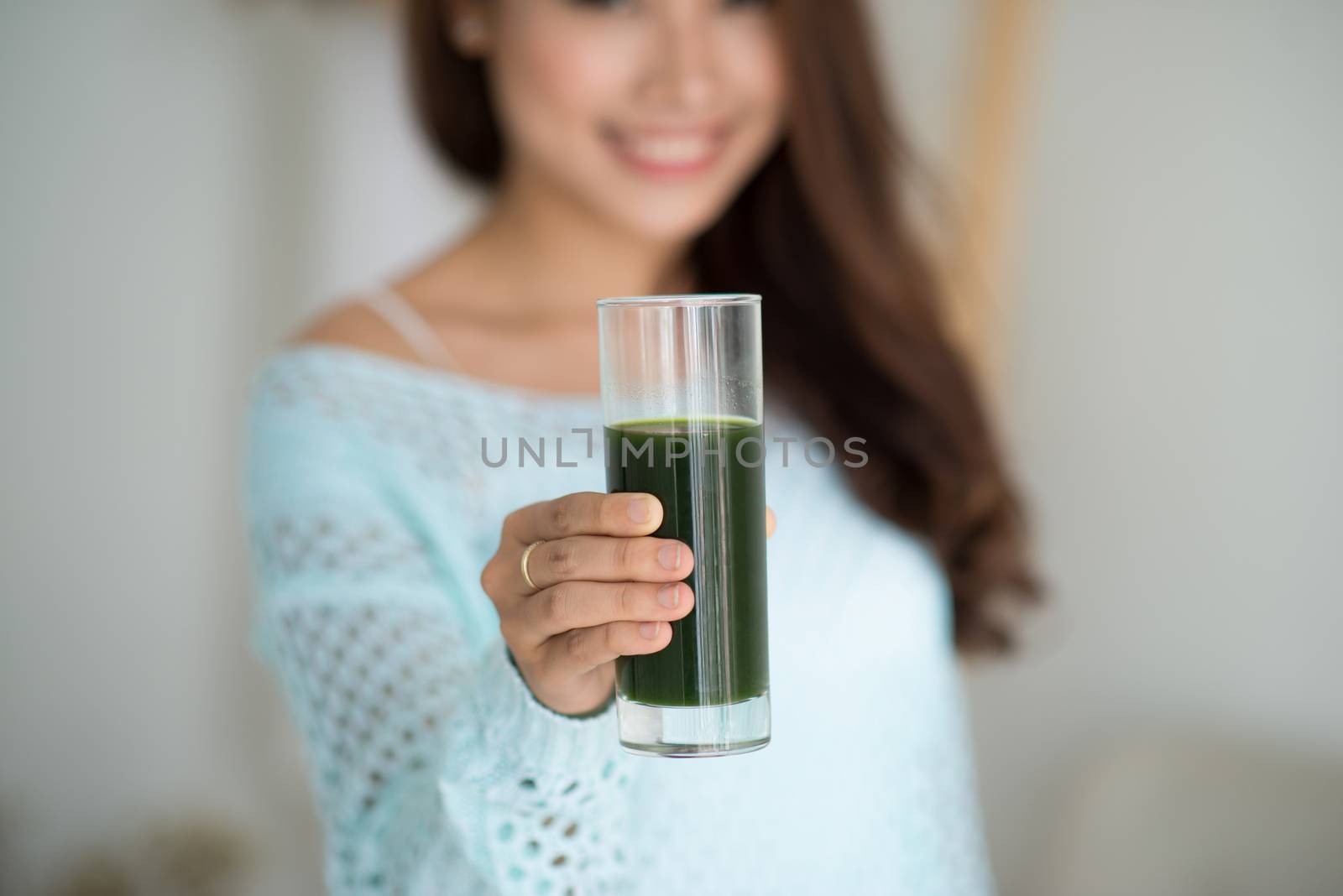 Pretty young asian woman drinking green fresh vegetable juice or smoothie from glass at home