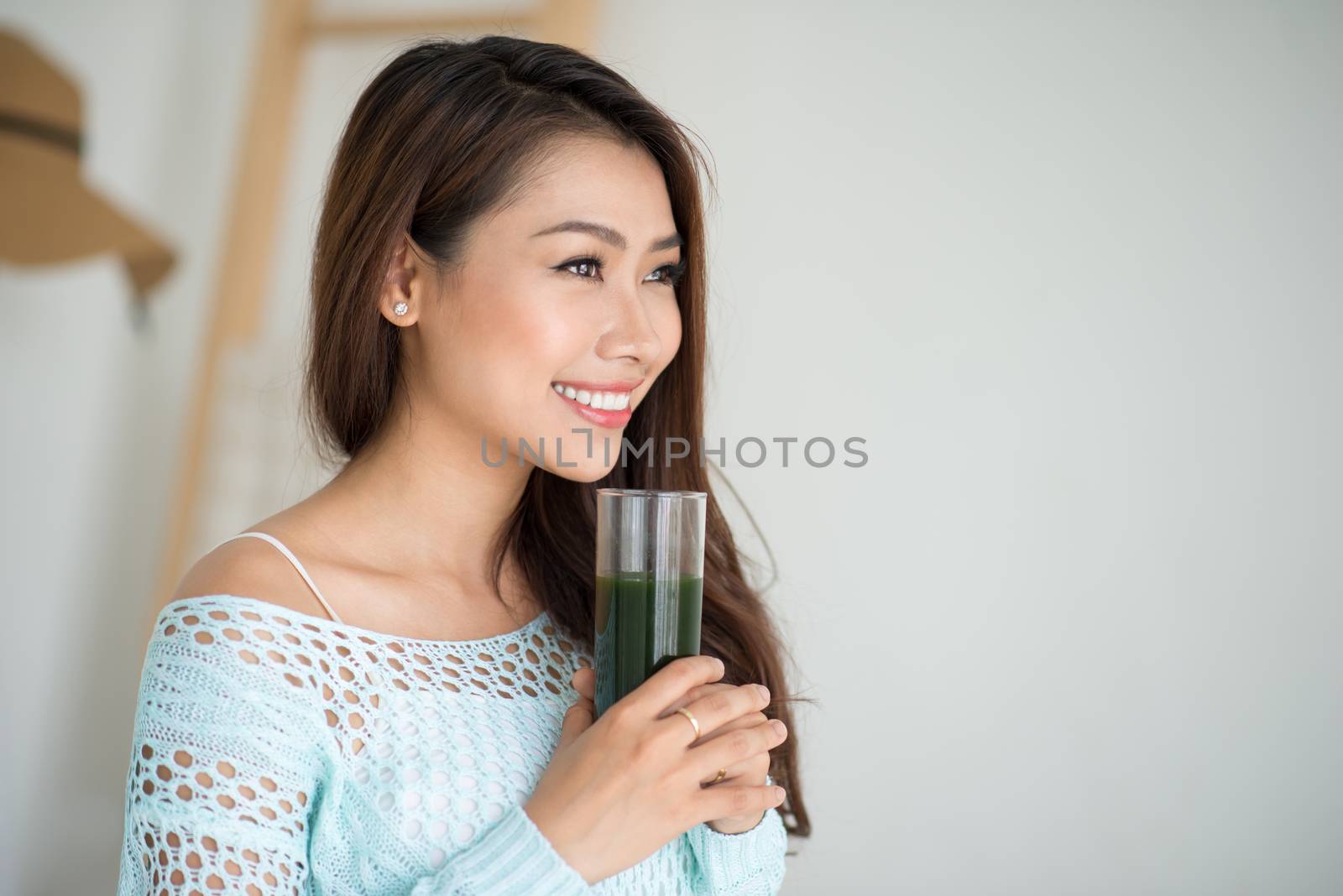 Pretty young asian woman drinking green fresh vegetable juice or smoothie from glass at home