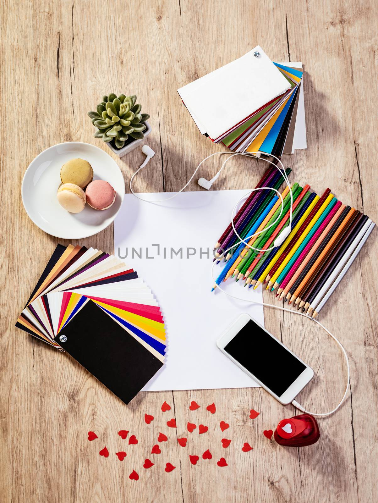 Top view of a designer workspace with blank paper and pallete color. Different office accessories, smartphone with blank screen and franch macaroons is on the table. 