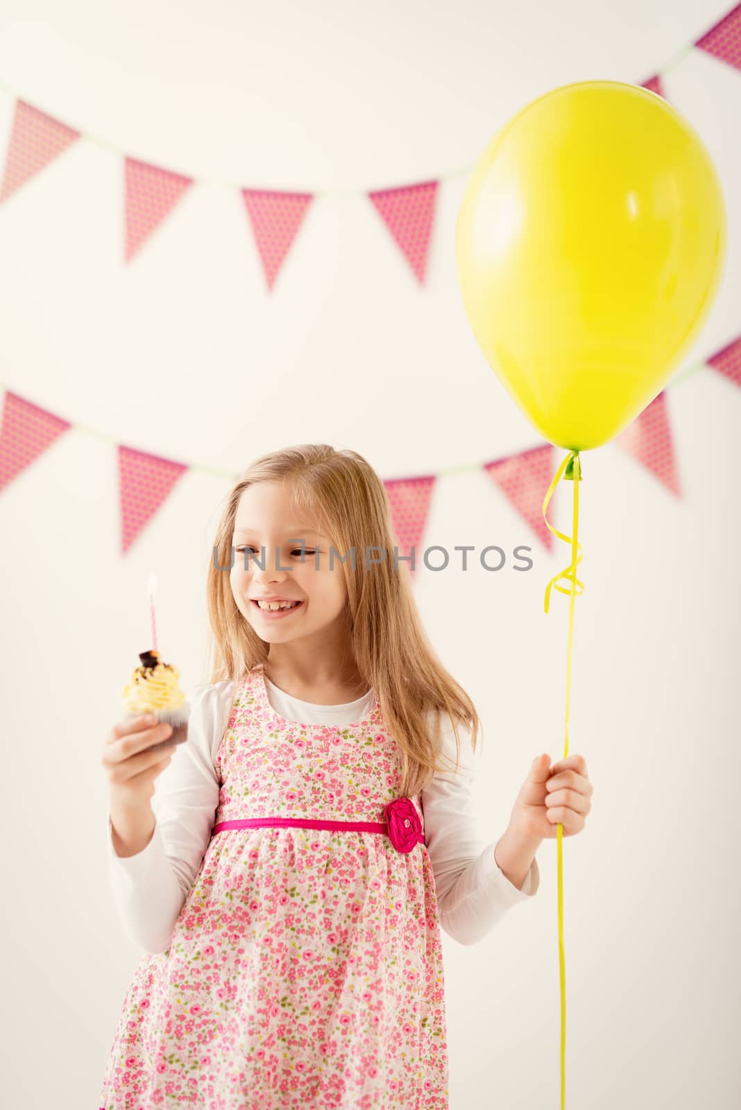 Beautiful cheerful little girl holding balloon and small birthday cake with candle.