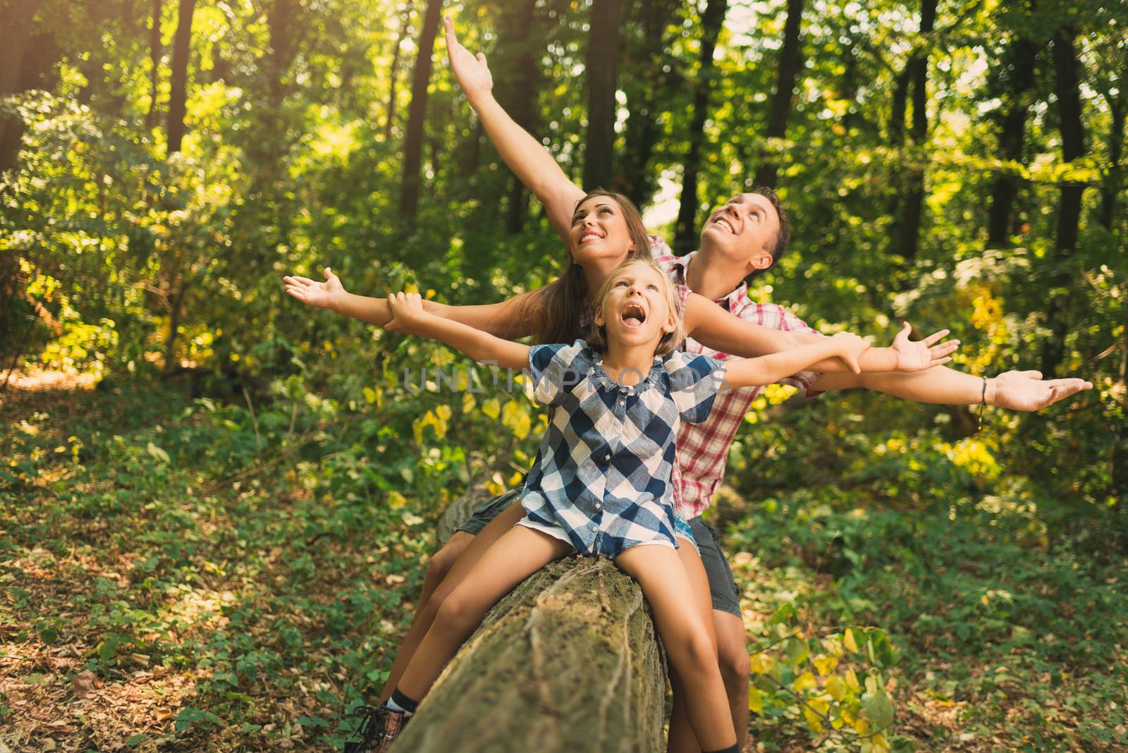 Young crazy family having fun during the walk through forest. They are sitting on fallen tree in the forest and looking up with open arms.