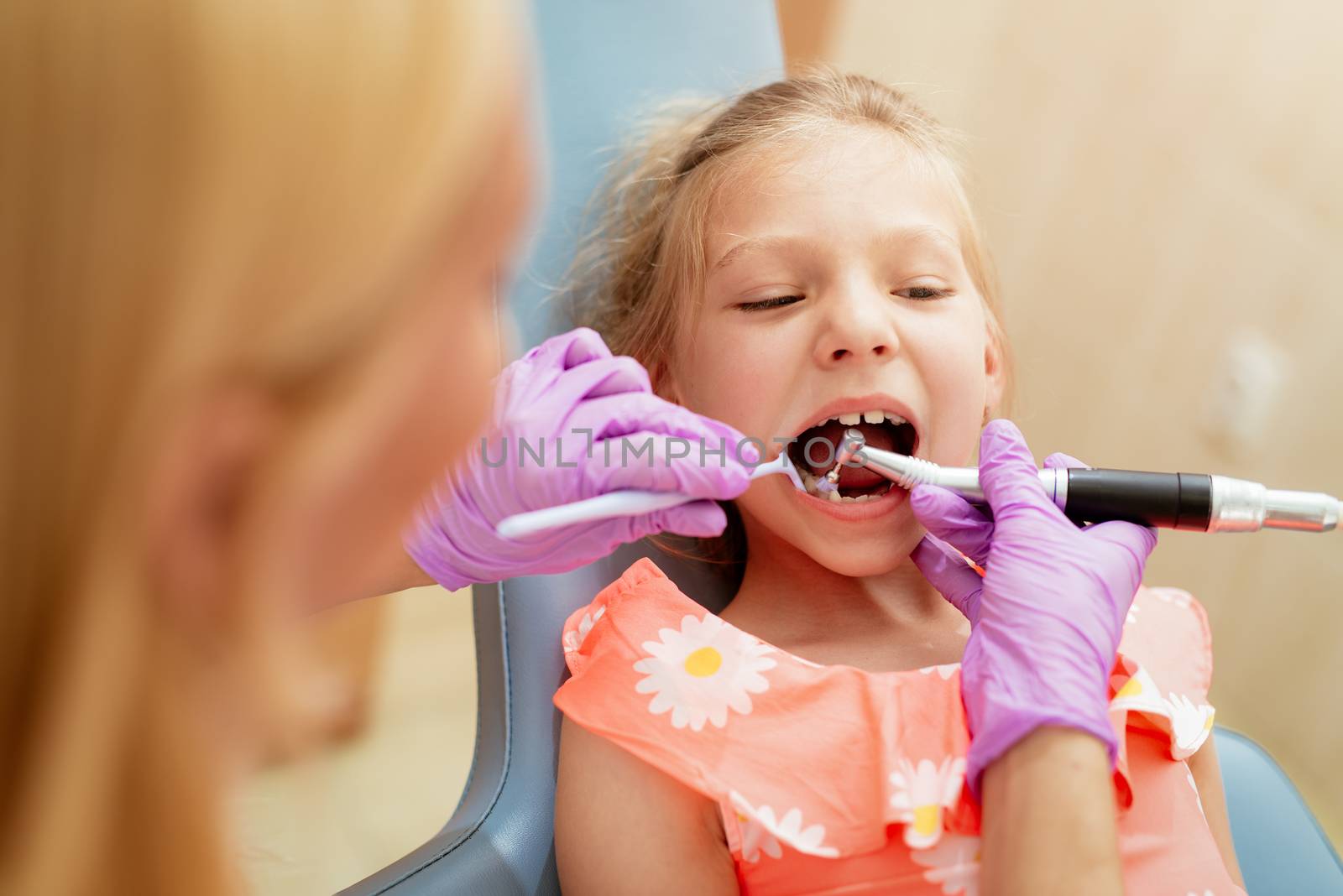 Beautiful little girl at visit in the dentist office. She is sitting on a chair and dentist polished teeth on her.