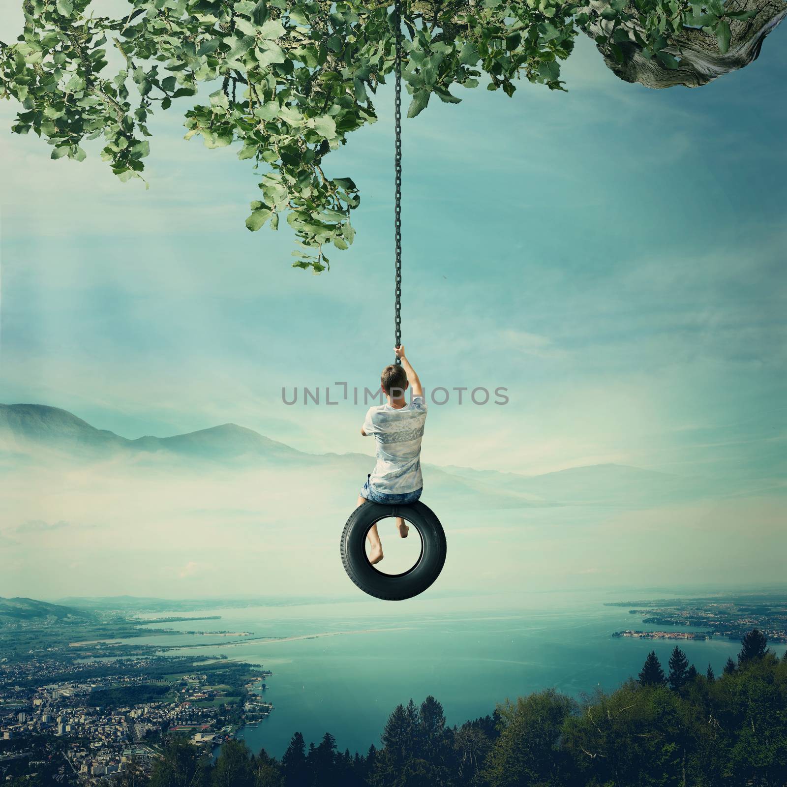 Young boy swinging on a tire over the foggy city with lake and forest background. Having fun and freedom concept