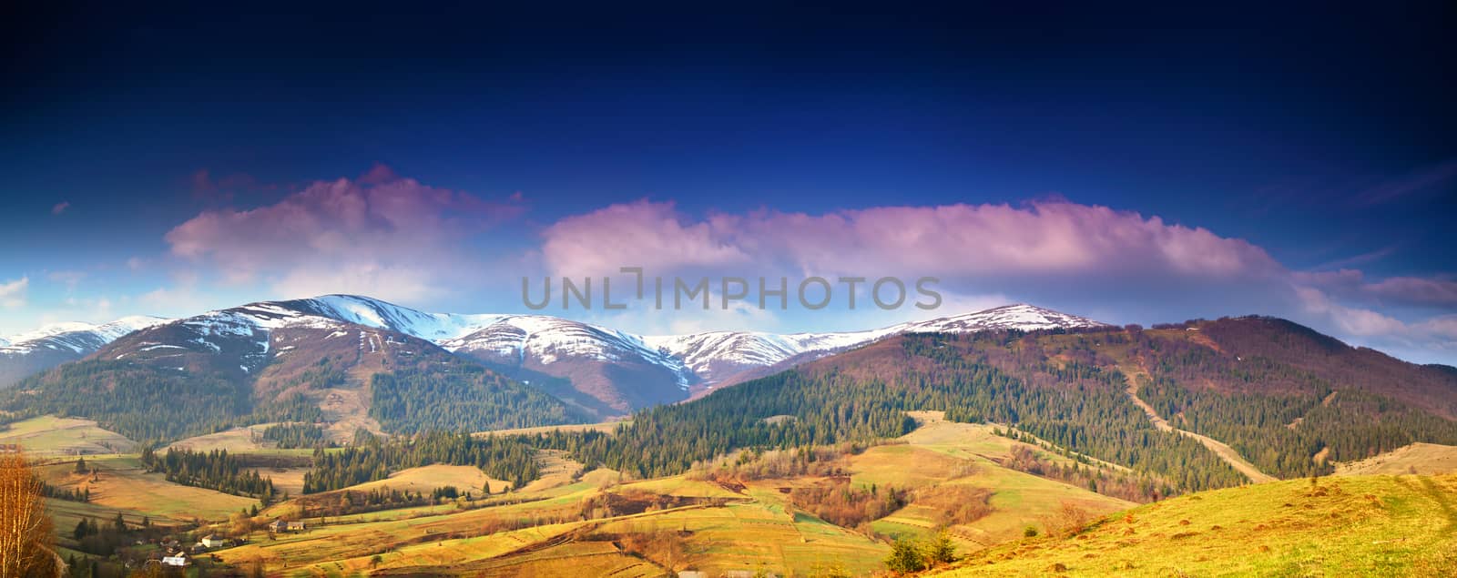 Panorama view of snow capped mountain peaks in spring time. Mountains in morning light. Deep blue sky. Idyllic Spring alpine morning landscape with fresh green meadows and snow-capped mountain tops in the background.
