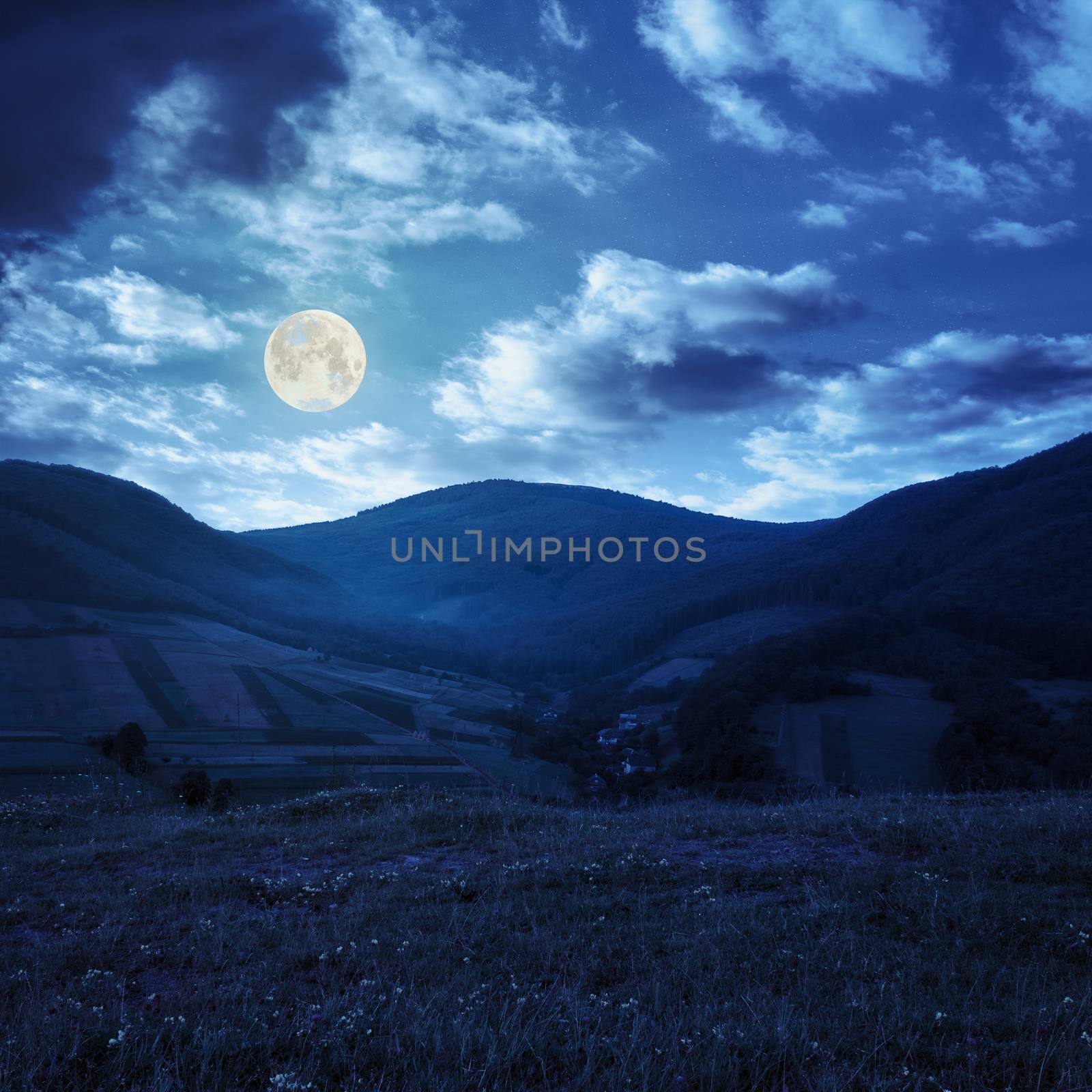 Stack of hay on a green meadow in the mountains in the morning under a blue summer sky at night in full moon light