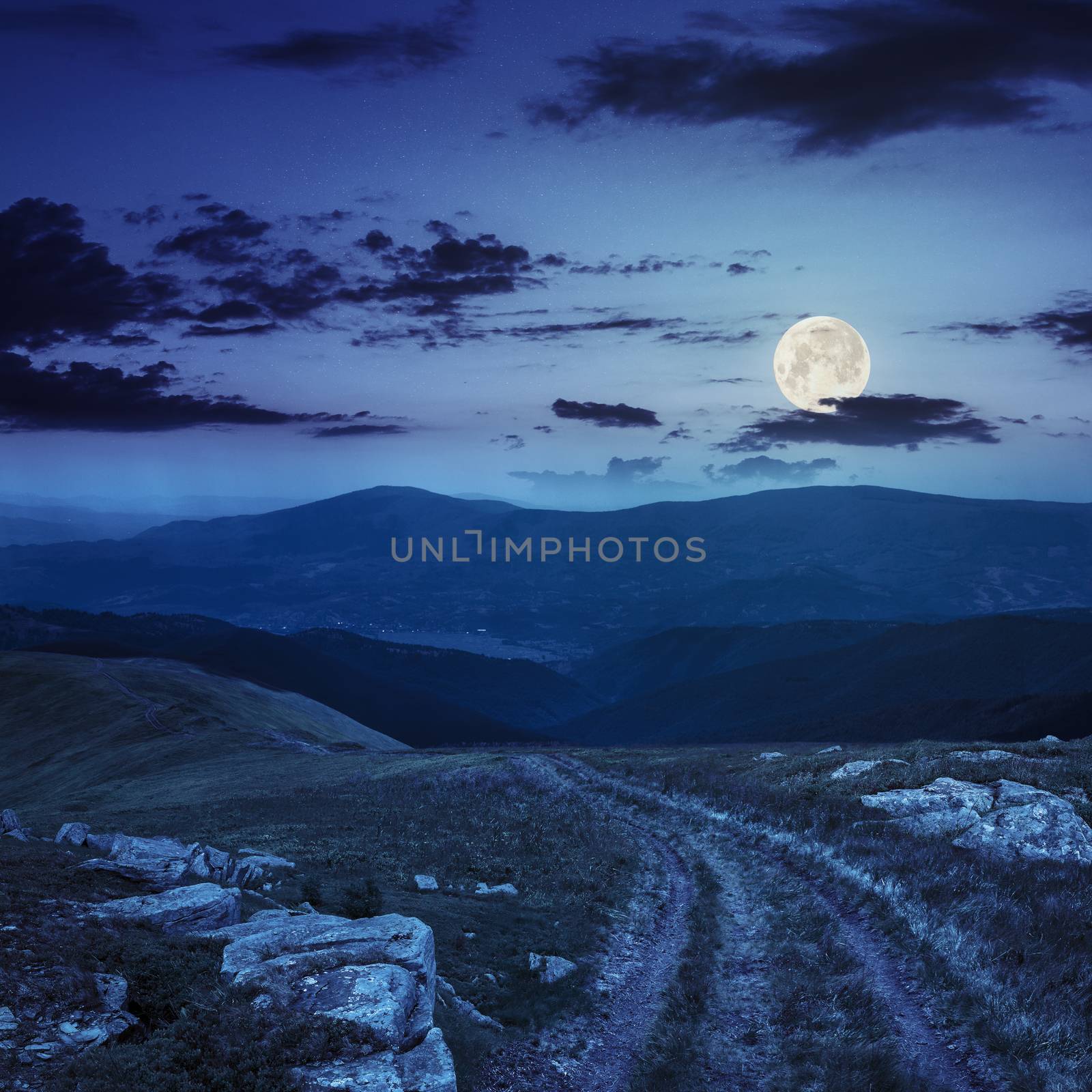 road among white sharp stones on the hillside on top of mountain range at night in full moon light
