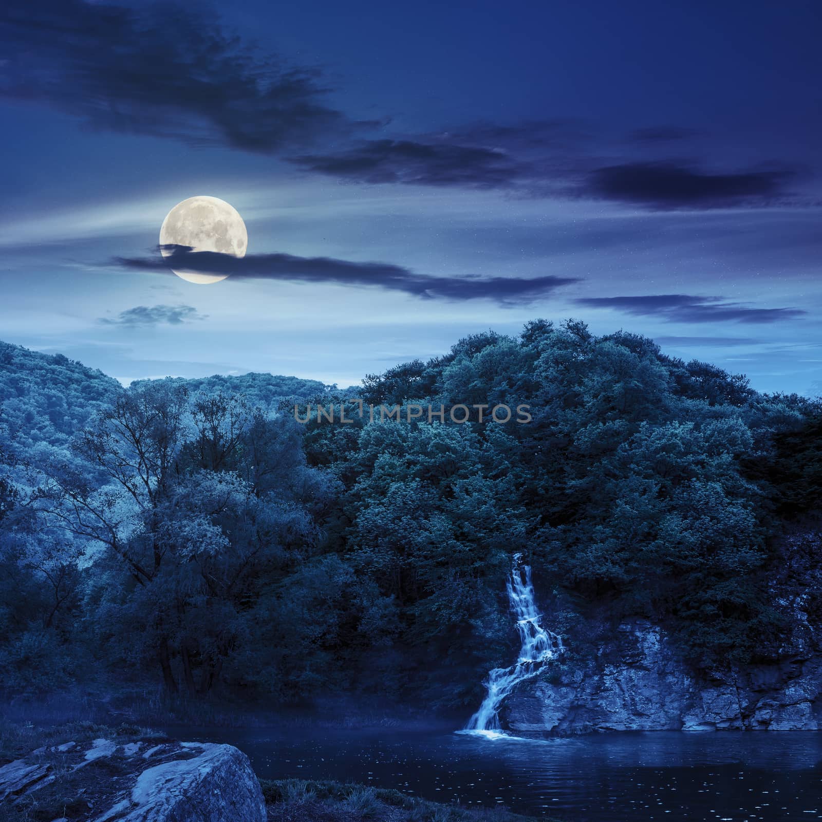 small waterfall comes out of a forest on a rocky hill and falls in river with fog at night in full moon light