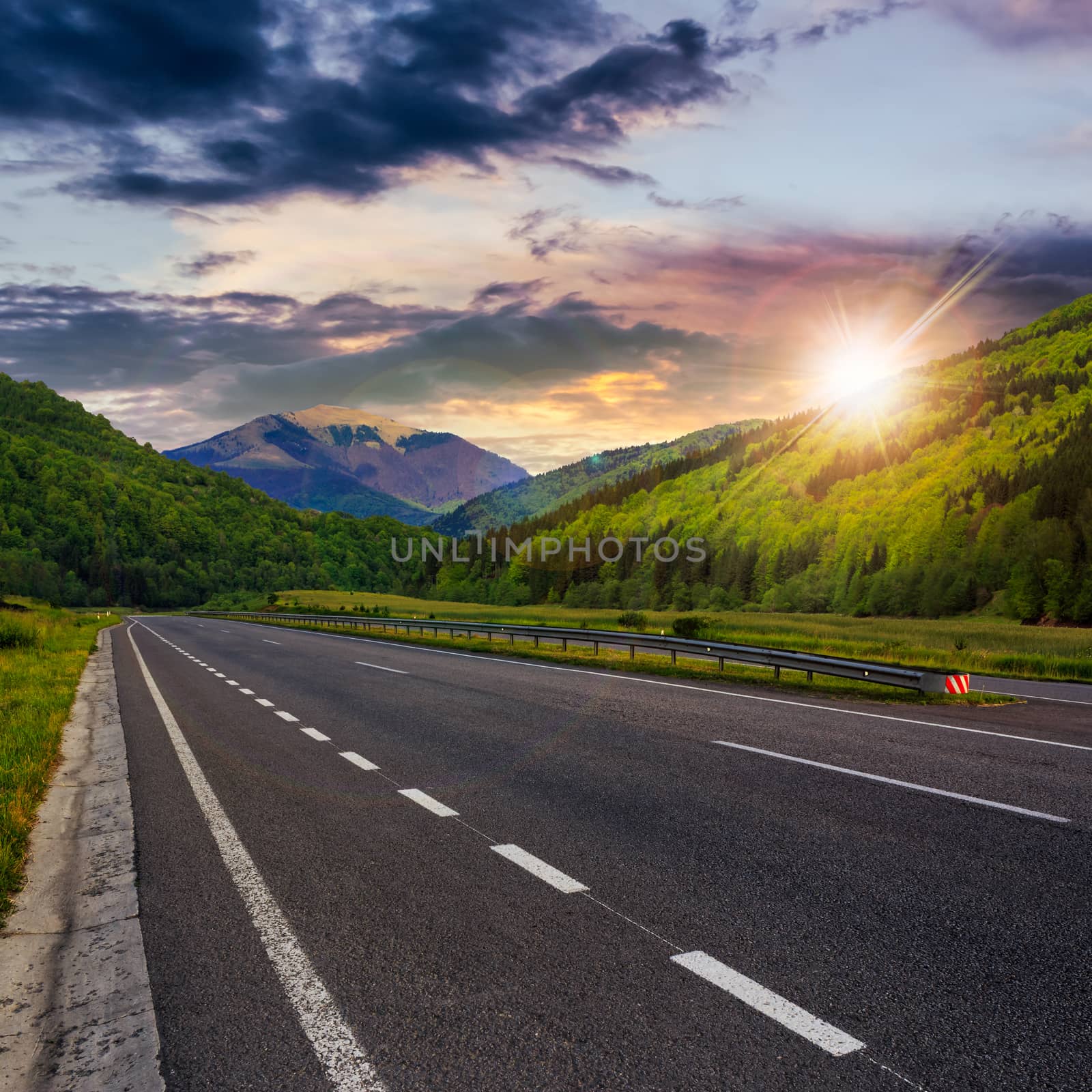 stright asphalt road in mountains passes through the green shaded forest at sunset