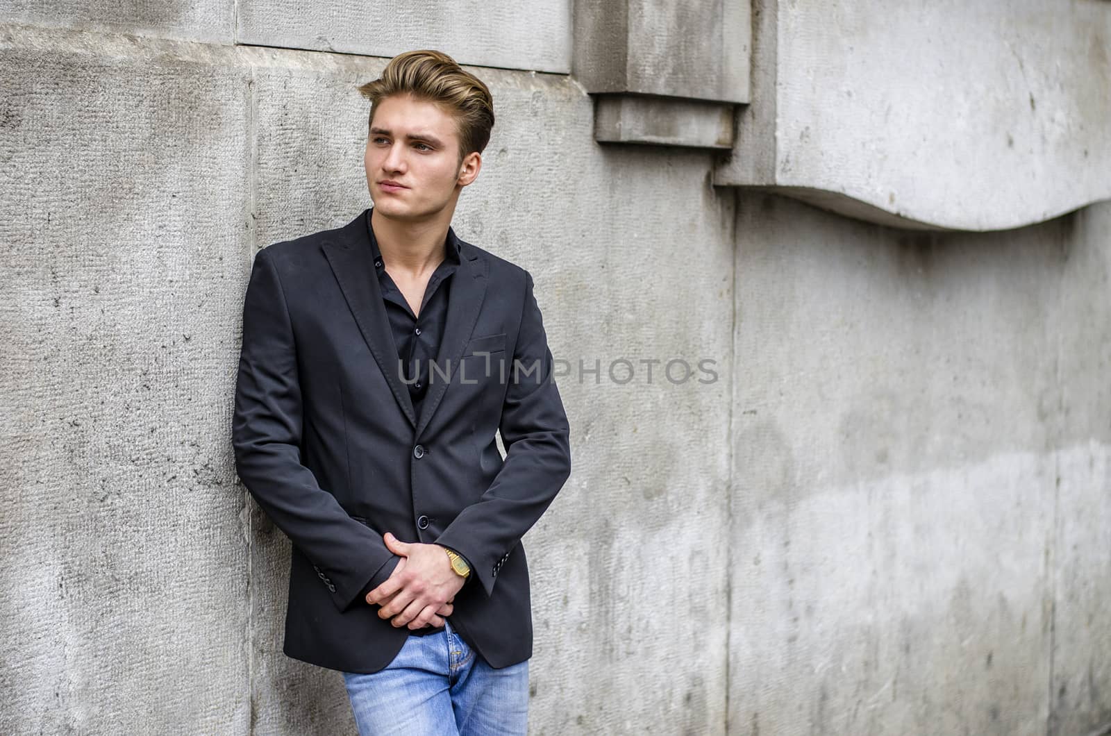 Handsome blond young man, blue eyes, leaning against white, rough stone wall, looking up to a side