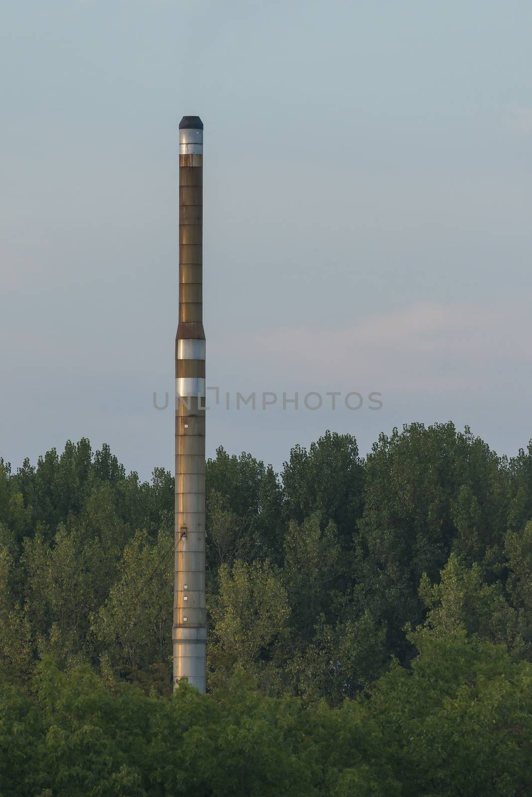 Factory chimney of a factory located in a forested area visible from a nearby higher point in the evening light
