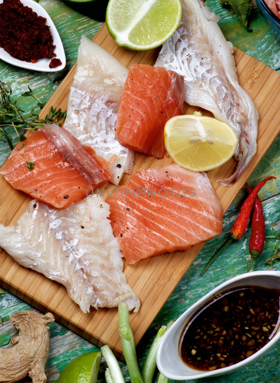 Arrangement of Raw Fillet of Salmon and Cod, Greens, Spices, Sauces and Lemon closeup on Cracked Wooden background