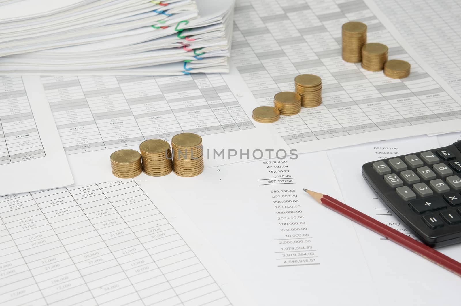 Brown pencil and calculator with step pile of gold coins on the statement finance account have blur overload of paperwork with colorful paperclip as background.