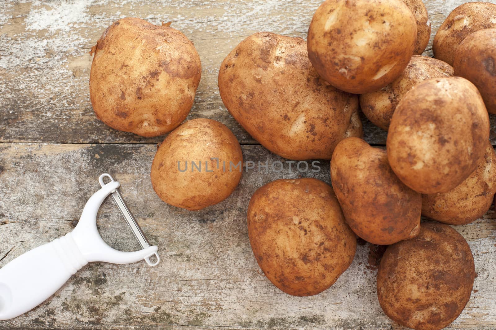 Pile of farm fresh potatoes still with adhering dirt and a manual peeler lying on a rustic background ready to prepare for dinner