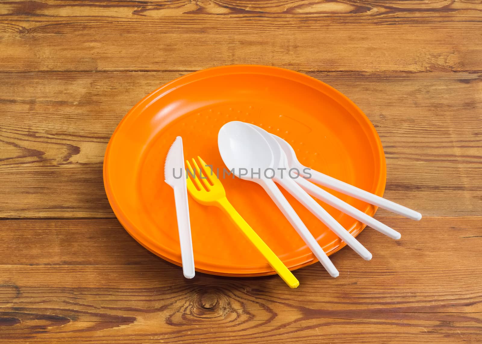 Several disposable plastic white spoons, knife, yellow fork on the pile of the orange plastic disposable plates on a surface of the old wooden planks
