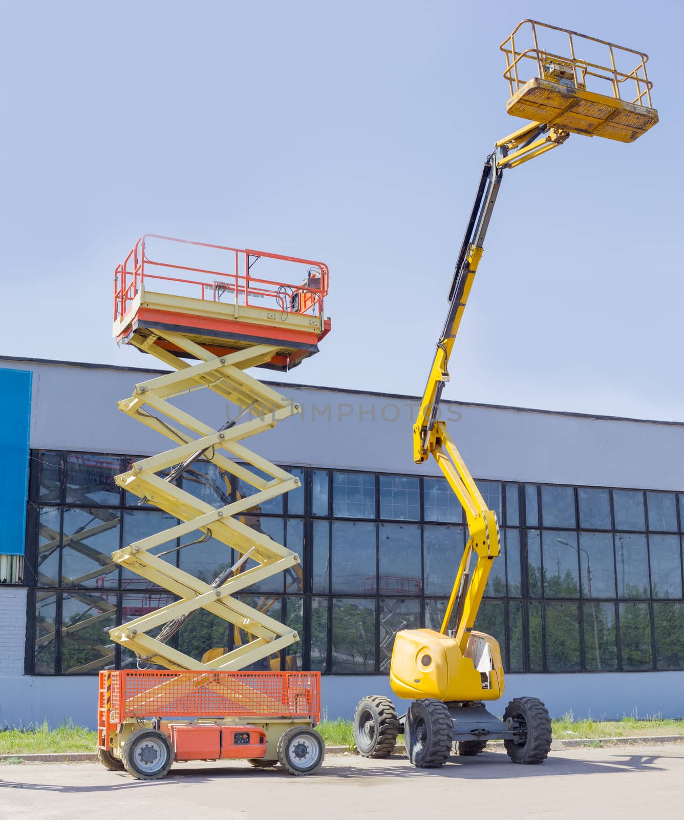 Wheeled scissor lift and wheeled articulated lift with telescoping boom and basket on an asphalt ground against the sky and an industrial building
