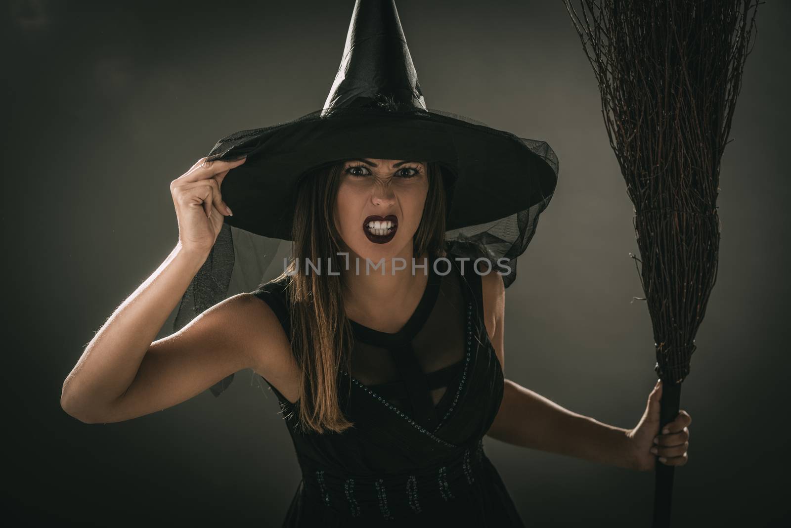 Portrait of a young woman dressed like a witch. She is in dark clothing and holding a broom. Looking at camera.