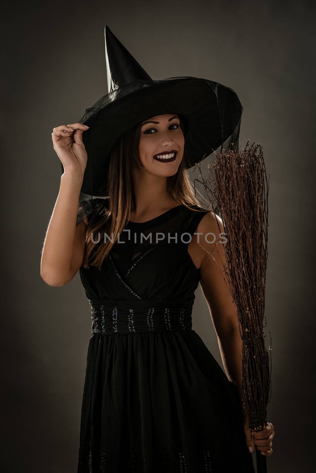 Young woman dressed like a witch. She is in dark clothing and holding a broom. Looking at camera.