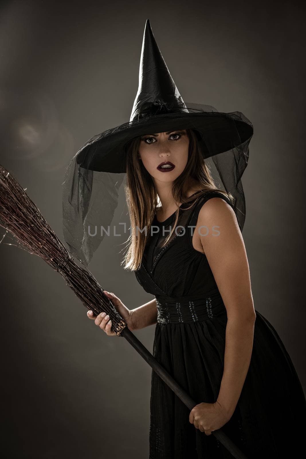 Young woman dressed like a witch. She is in dark clothing and holding a broom. Looking at camera.