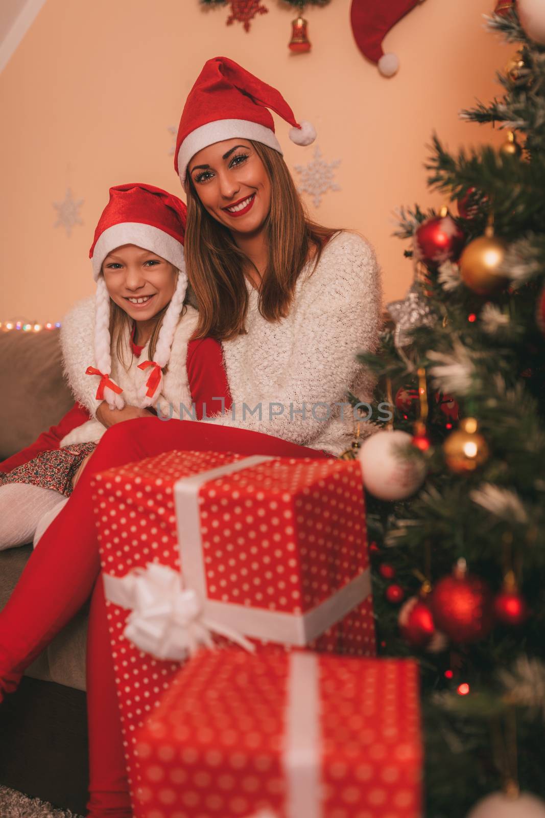 Cute smiling girl and her mother sitting next to a Christmas tree and many gift and looking at camera. They are at the home. 