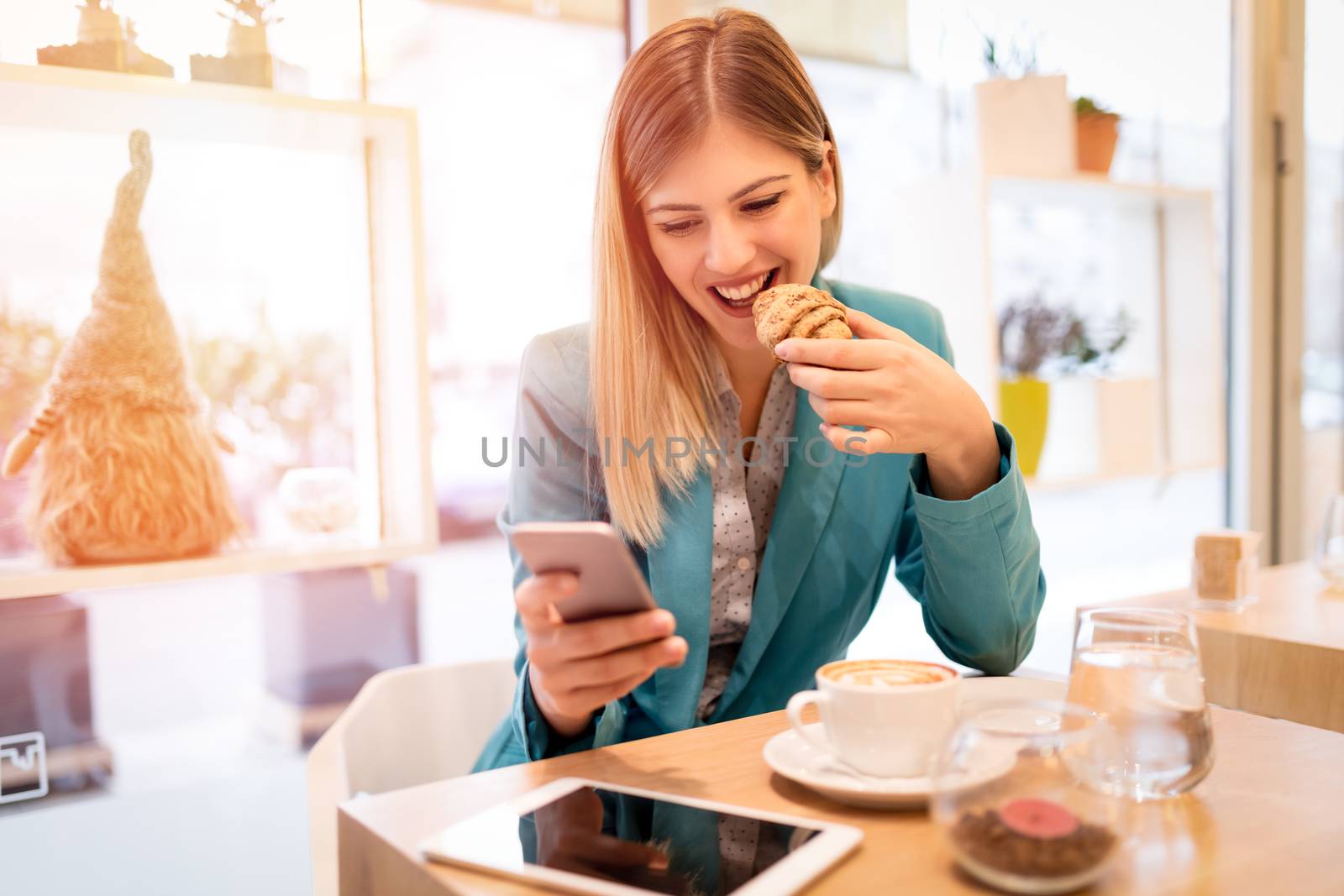 Young smiling businesswoman on a break in a cafe. He is eating croissant and reading message on smart phone. 
