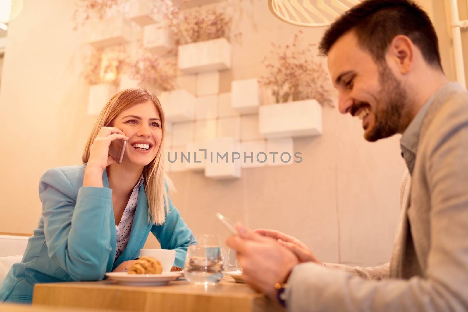 Two young smiling businesspeople on a break in a cafe. Man working at tablet. Woman using smart phone. They are drinking coffee and having breakfast. Selective focus.