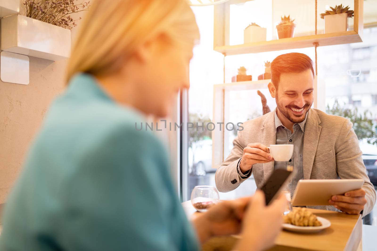 Young smiling businesspeople on a break in a cafe. Man working at tablet and drinking coffee. Woman using smart phone. Selective focus. Focus on background, on businessman.