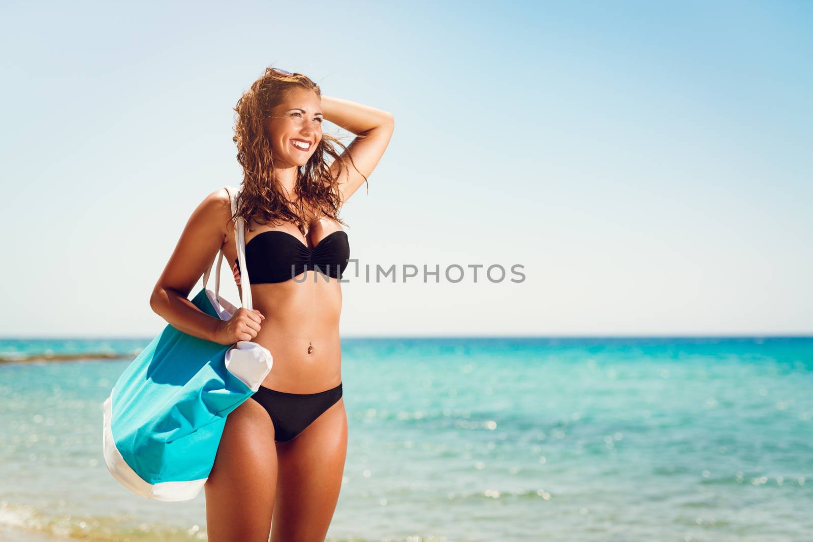 Portrait of a beautiful young woman enjoying on the beach. She is holding summer bag and looking away with smiling on her face.