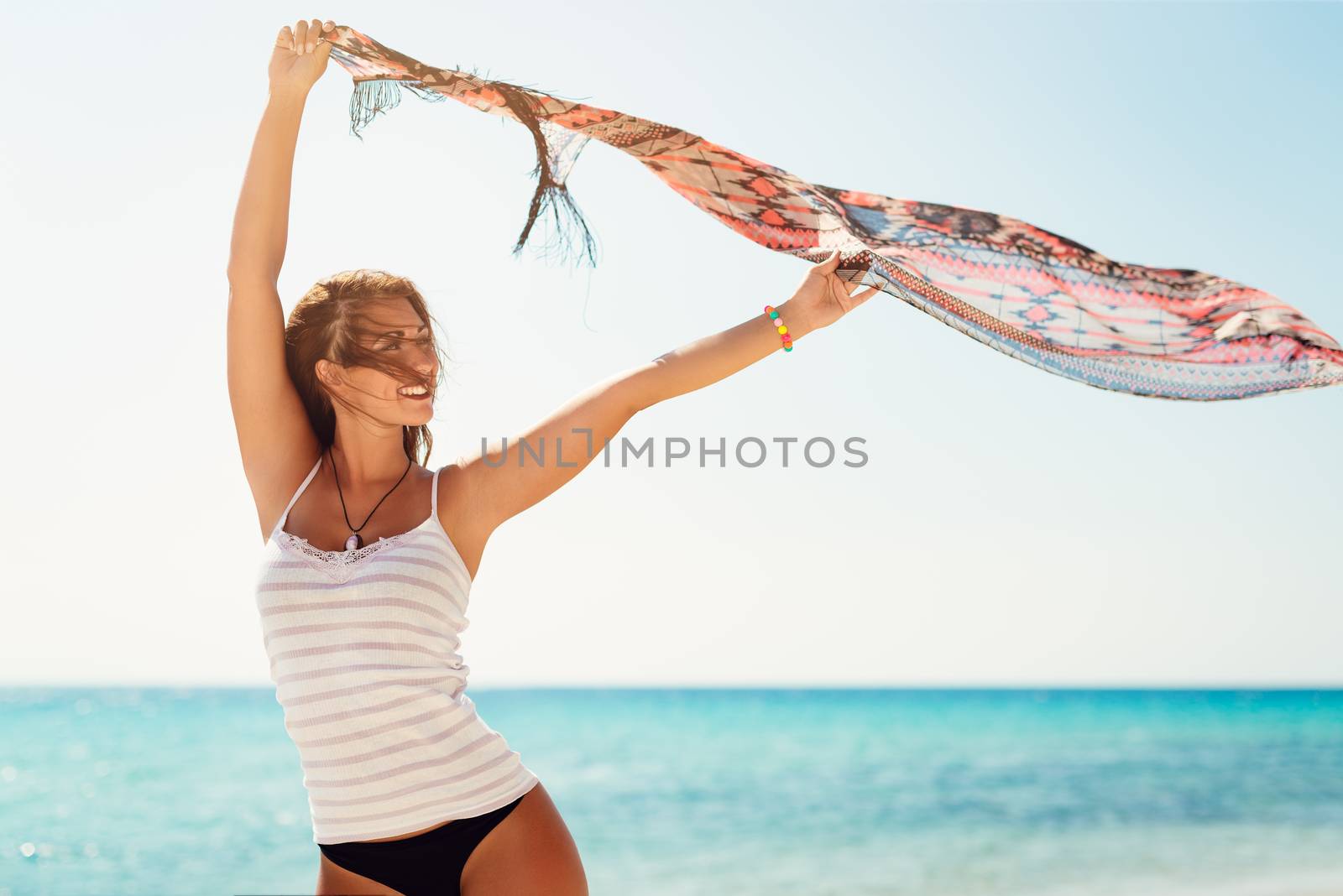 Beautiful young woman posing on the beach. She is holding scarf and looking away with smiling on her face.