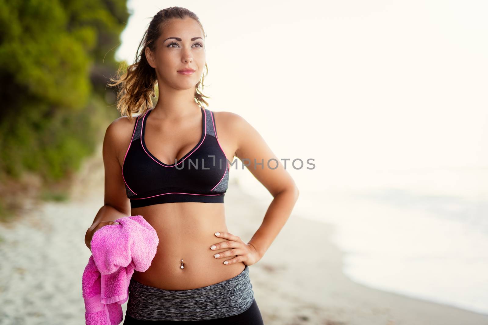 Beautiful young woman relaxing after training on the sands beach. She is holding towel and looking away.