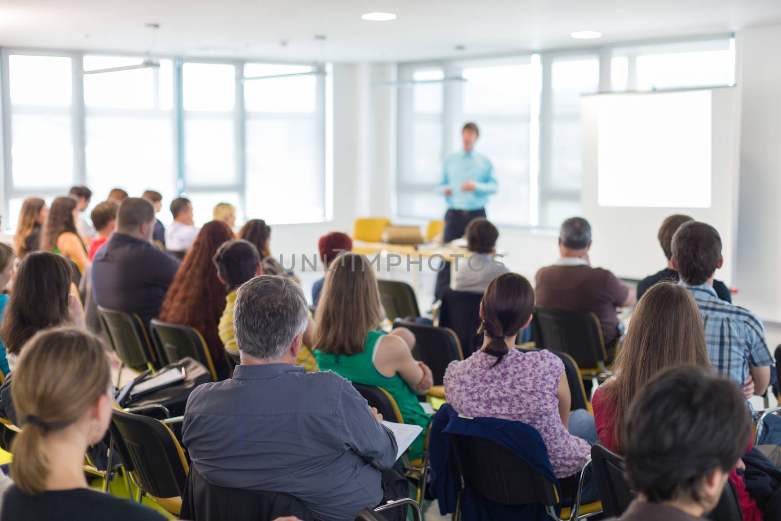 Business and entrepreneurship symposium. Speaker giving a talk at business meeting. Audience in conference hall. Rear view of unrecognized participant in audience.