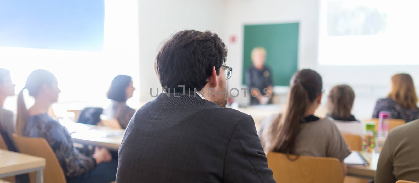 Speaker giving presentation in lecture hall at university. Participants listening to lecture and making notes.