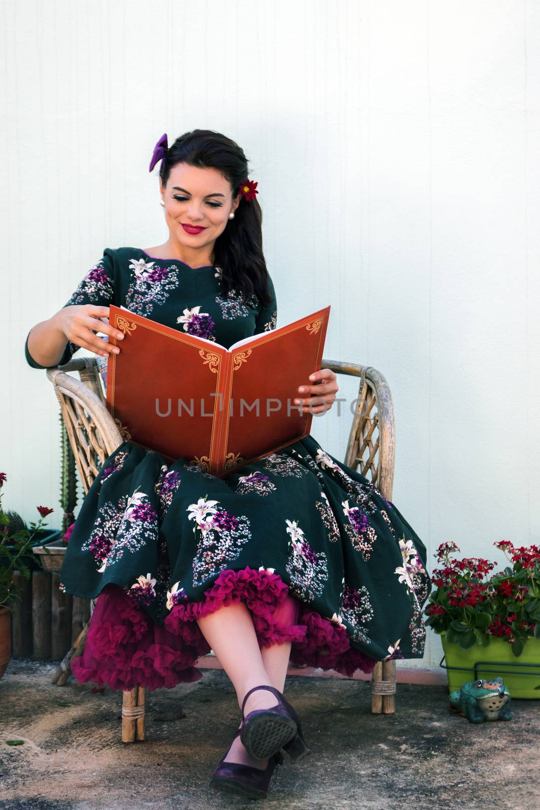 Vintage girl with beautiful floral dress reading a book on a wicker chair.