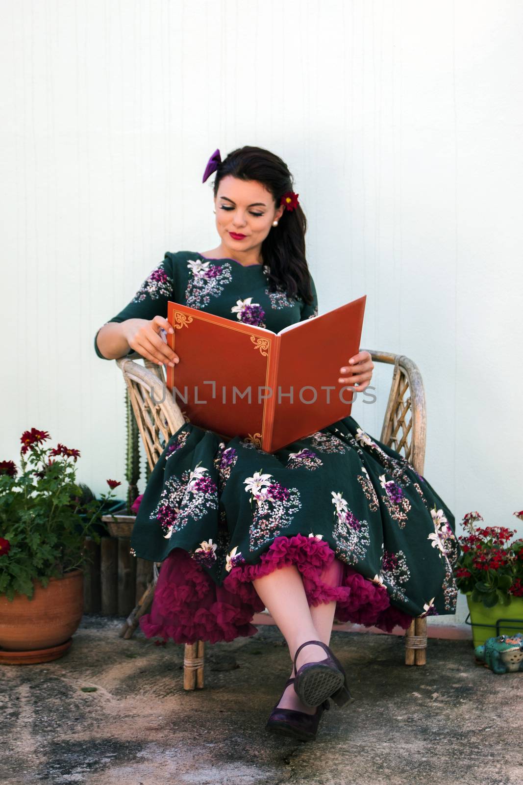 Vintage girl with beautiful floral dress reading a book on a wicker chair.