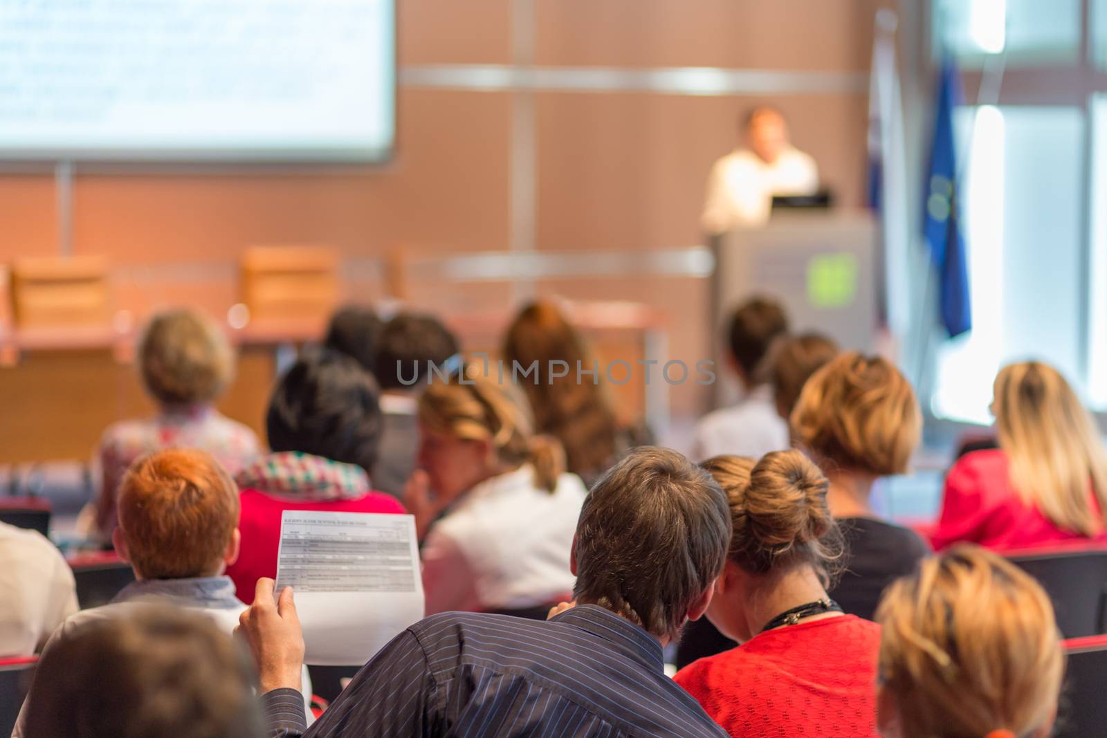 Business and entrepreneurship symposium. Speaker giving a talk at business meeting. Audience in conference hall. Rear view of unrecognized participant in audience.