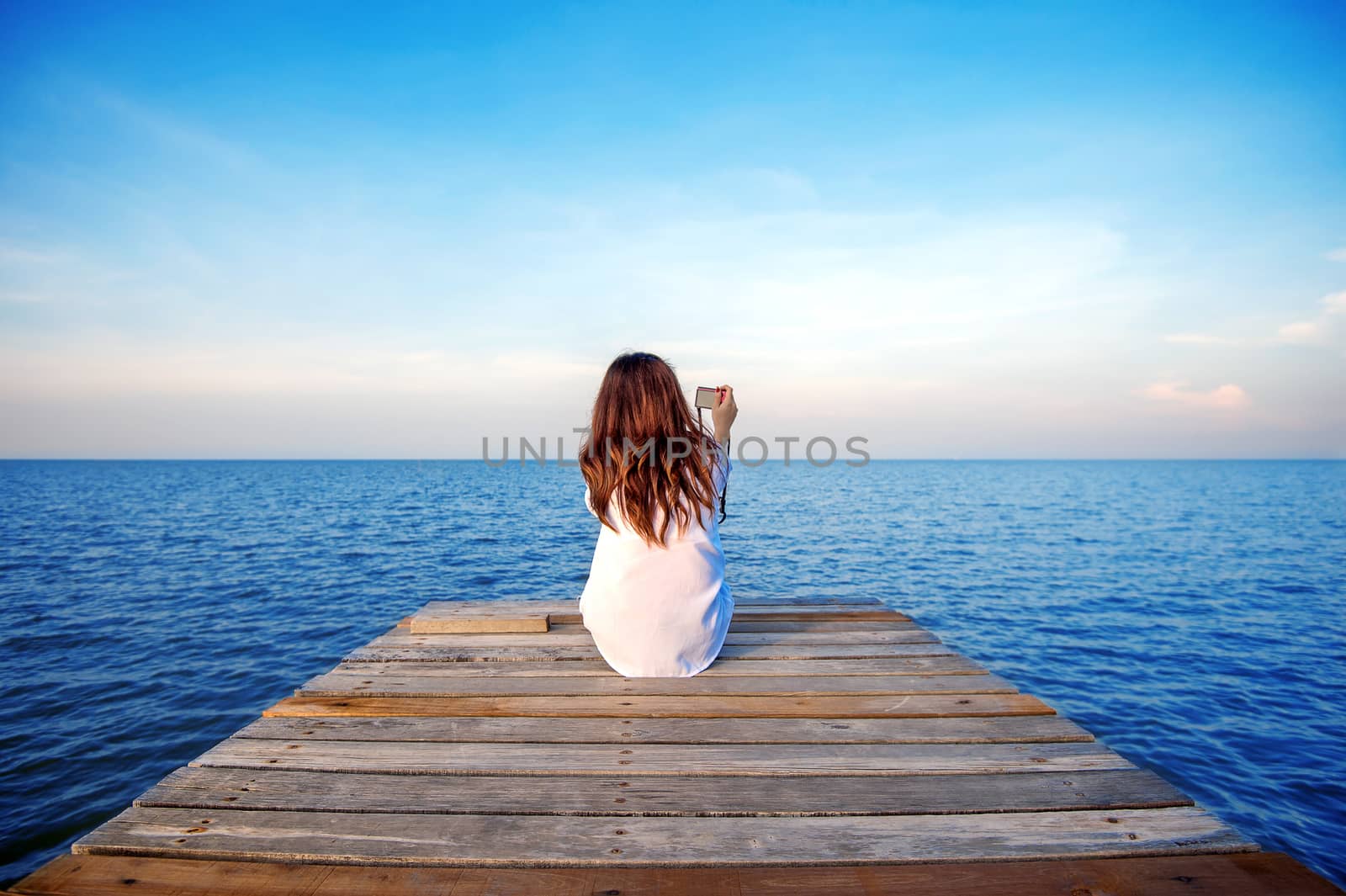 Girl sitting alone and hand holding camera on a the wooden bridge on the sea.