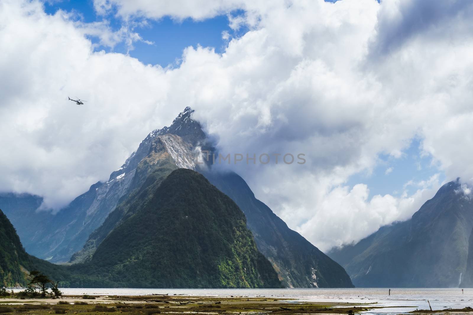 Helicopter flying over the valleys of the  Milford Sound. Helicopter dwarfed by the size of Mitre Peak mountain in the background shrouded by clouds.