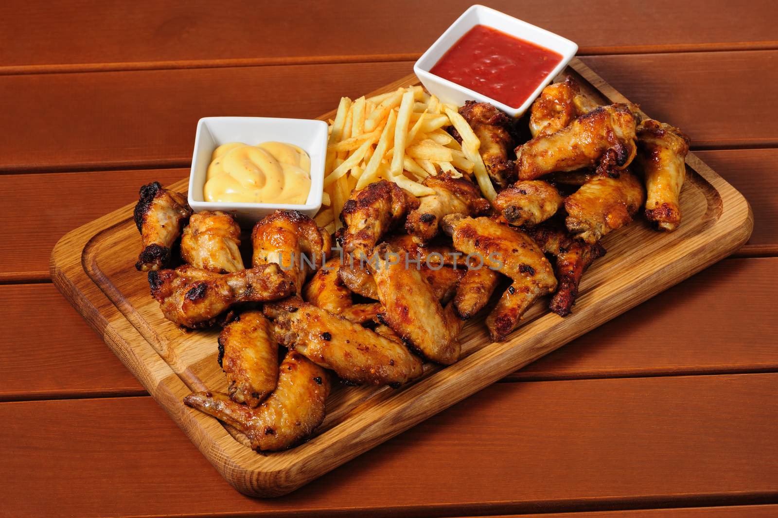 big wooden board with grilled chicken wings with french fries and two sauces, preferably served as companion for beer or other alcohol drinks