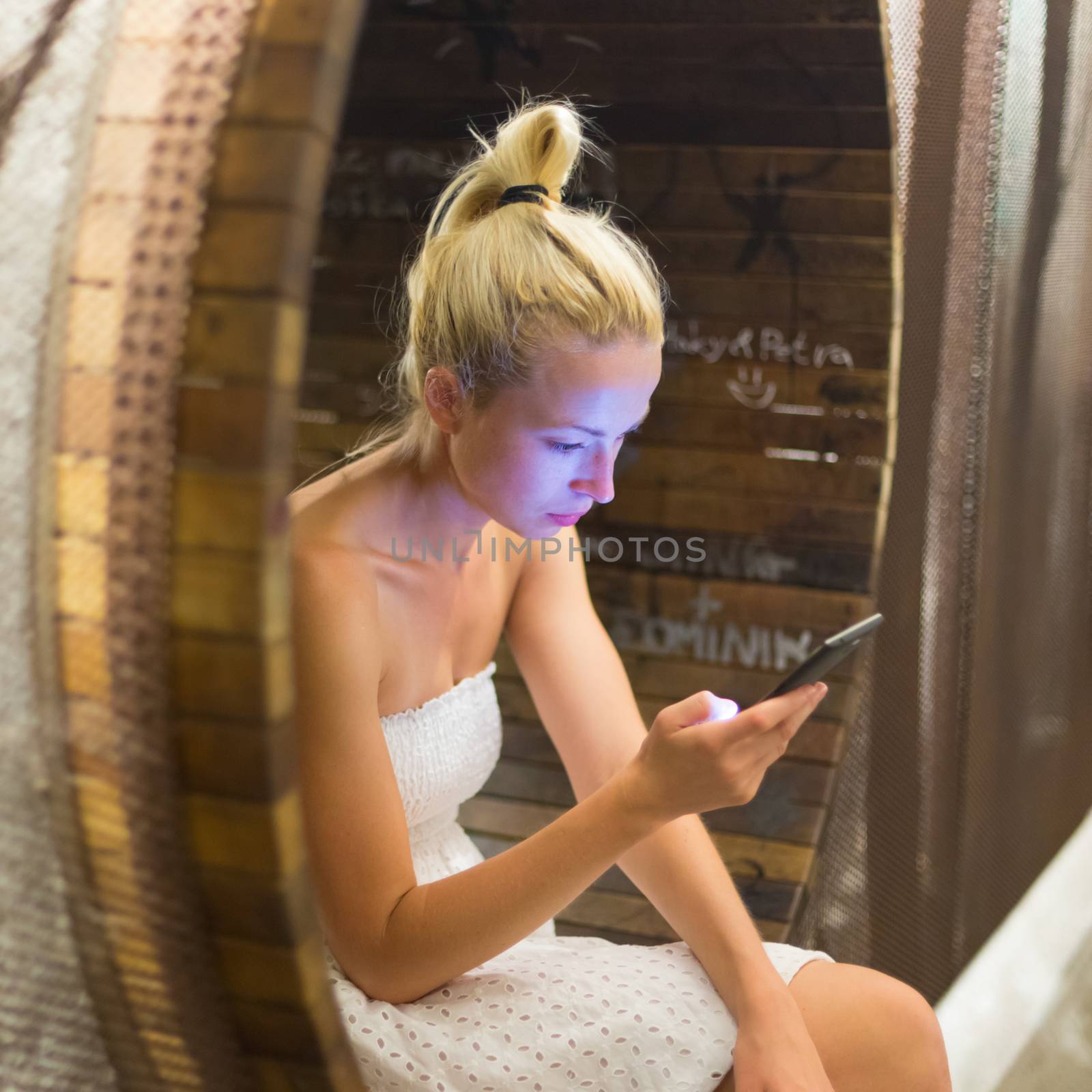 Casual young lady wearing white summer dress playing carelessly with her android smarthphone browsing trough social networks sitting on contemporary circular bench in city park.