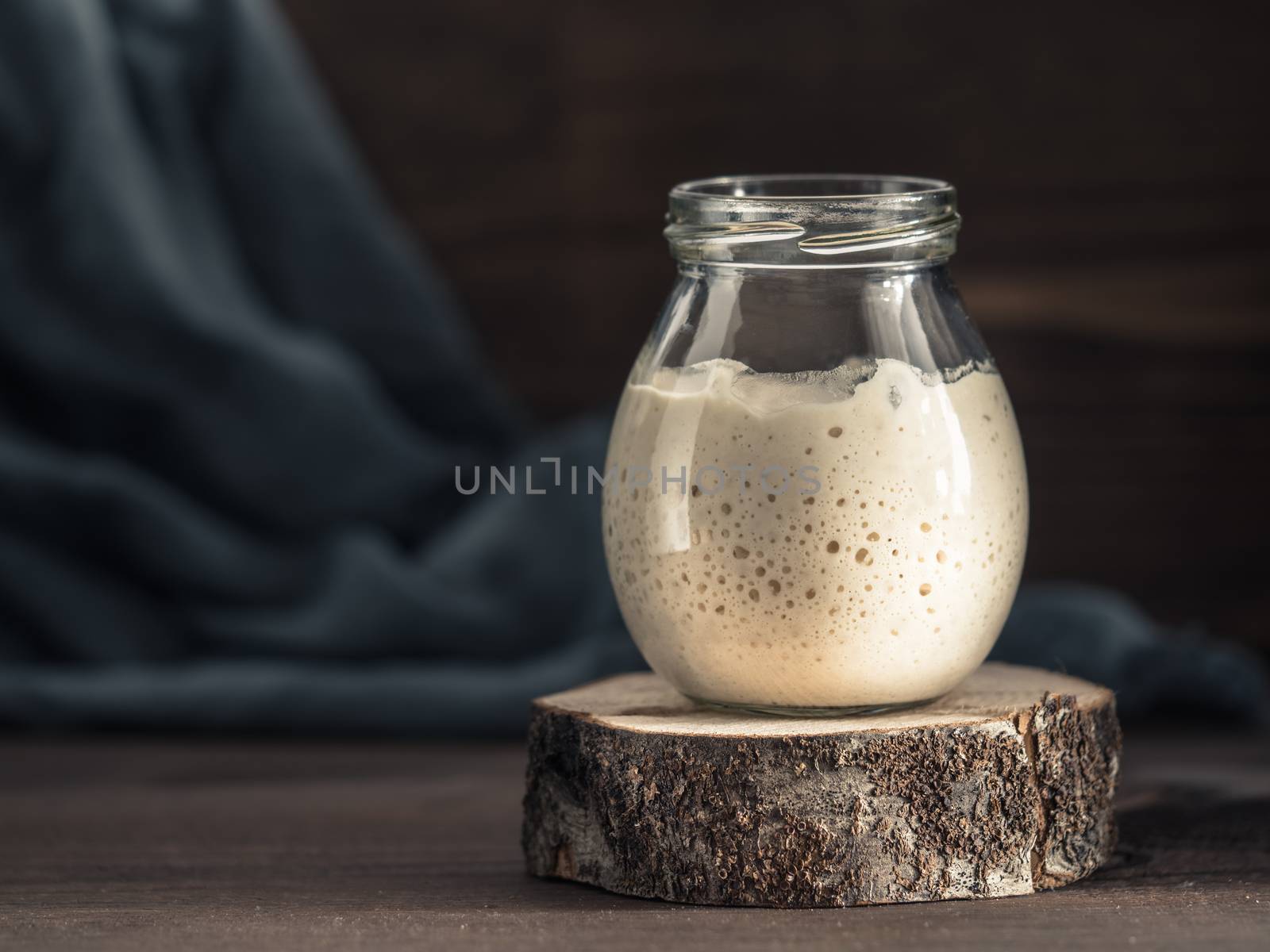 Active wheat sourdough starter in glass jar on brown wooden background. Starter for sourdough bread. Toned image. Copy space.