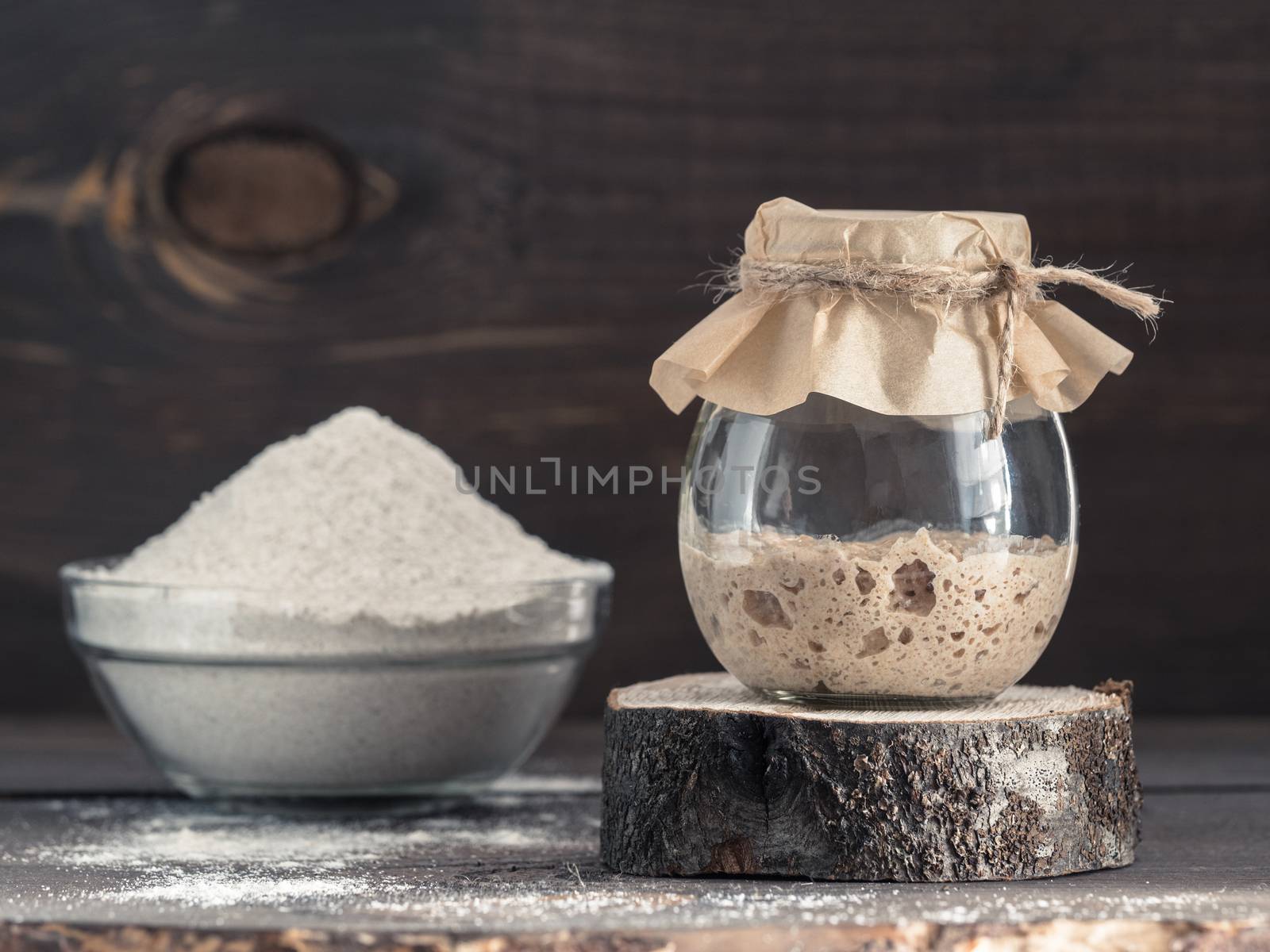 Active rye sourdough starter in glass jar and rye flour on brown wooden background. Starter for sourdough bread. Toned image. Copy space.