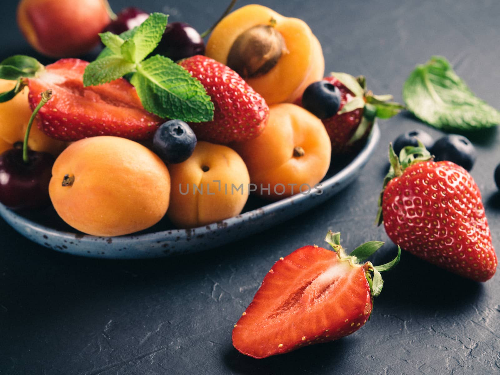 Closeup view of fruits and berries on dark background. Heap of fresh strawberries, blueberries, apricot and mint leaves in trendy plate. Focus on strawberry. Healthy food,superfood,diet,detox concept.