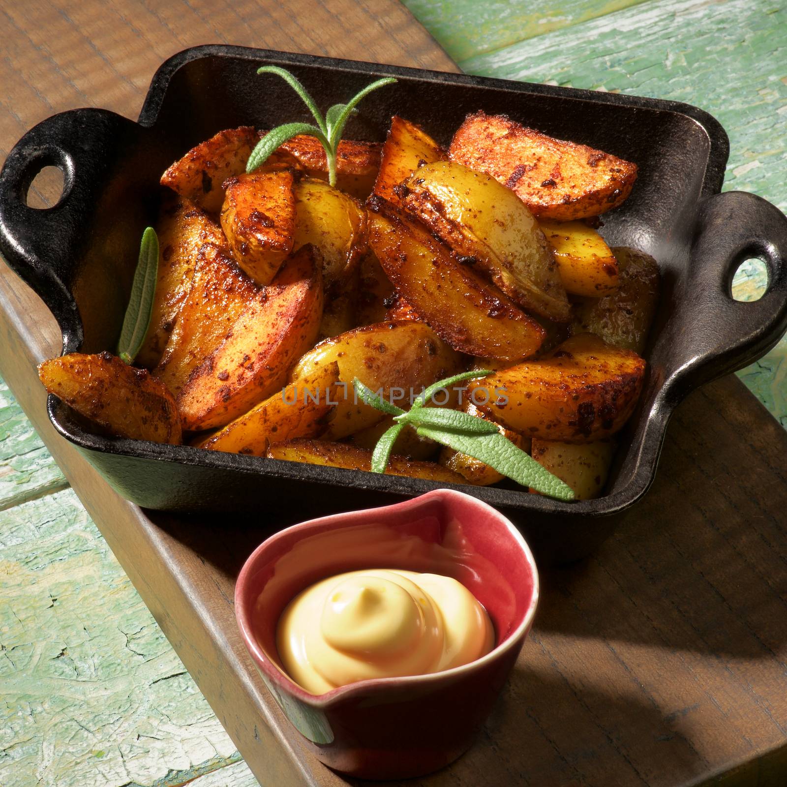 Hot Snack with Delicious Roasted Potato Wedges in Black Cast-Iron Pan and Cheese Sauce on Wooden Board closeup in Shadow
