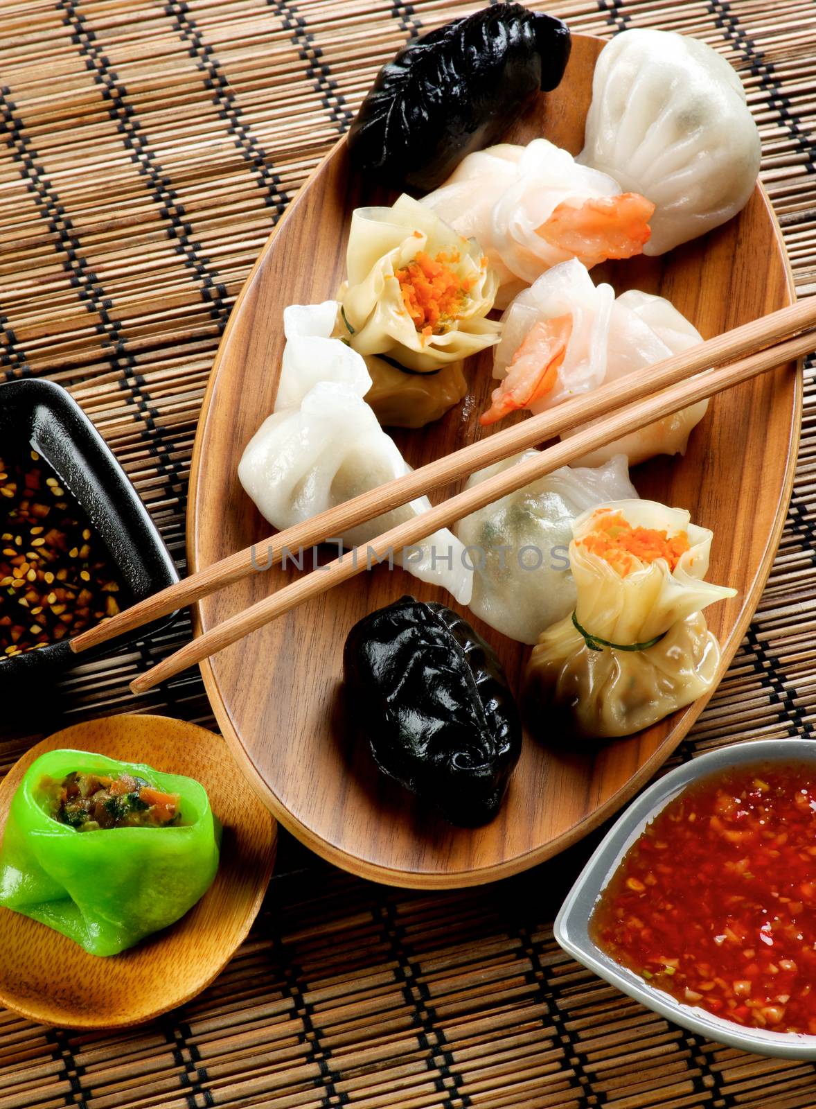 Various Dim Sum on Wooden Plate, Red Chili and Soy Sauces and Chopsticks closeup on Straw Mat background