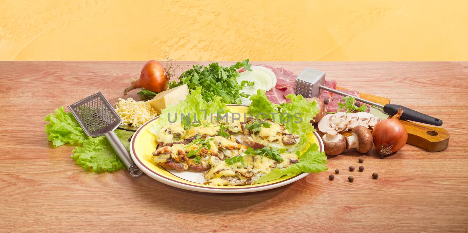 Pork chops baked with onion, mushrooms and cheese on the dish on the background of the ingredients and several kitchen utensils for preparation of chops on a wooden surface
